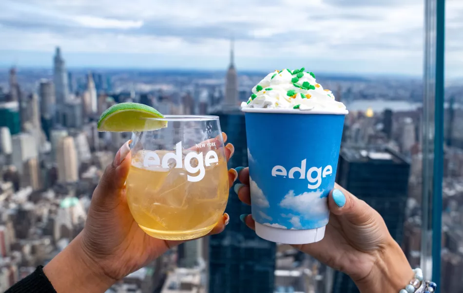 Two people clink glasses with festive St Patrick's day drinks overlooking New York city on a sunny day