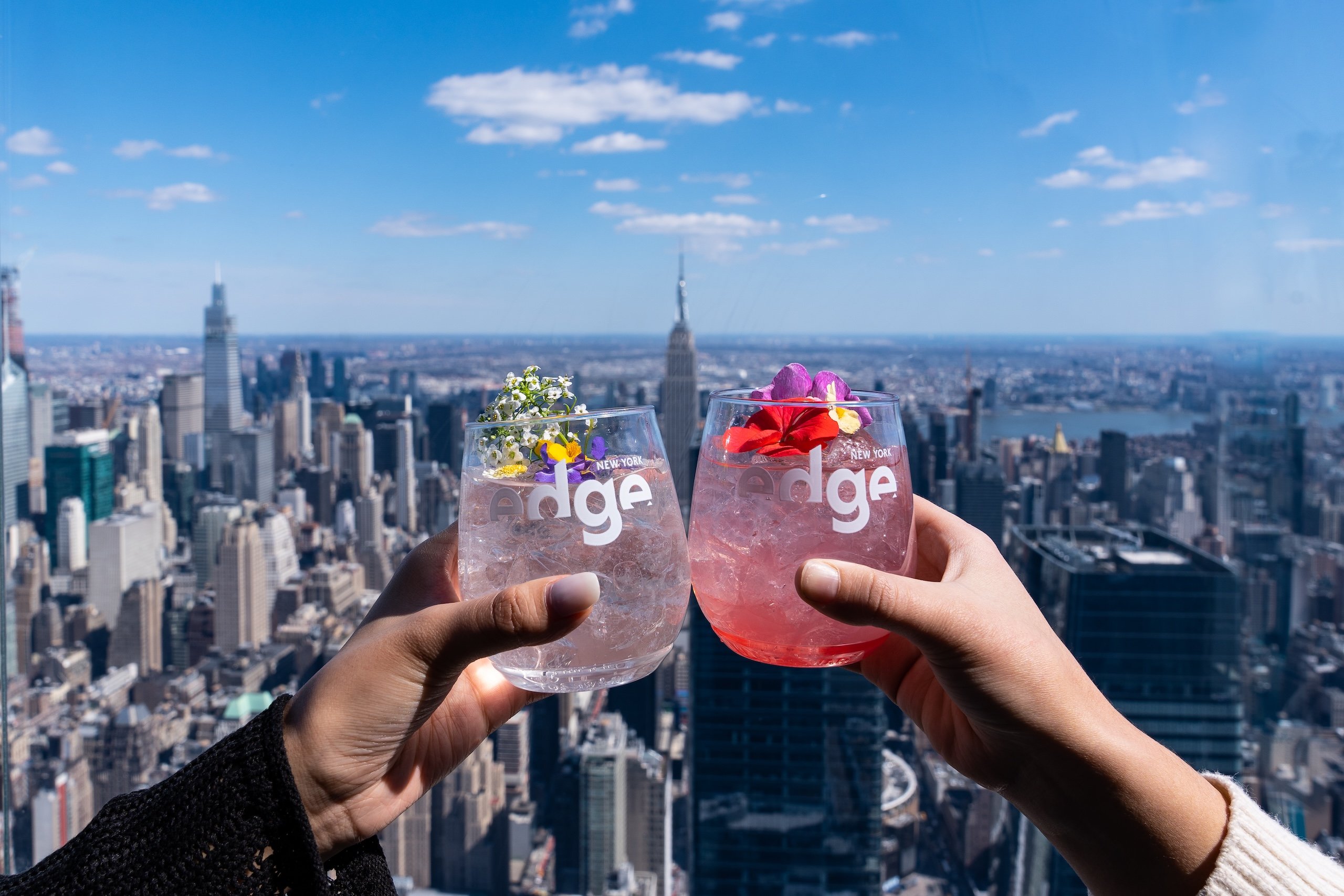 Two people clink glasses with cocktails overlooking New York city on a sunny day