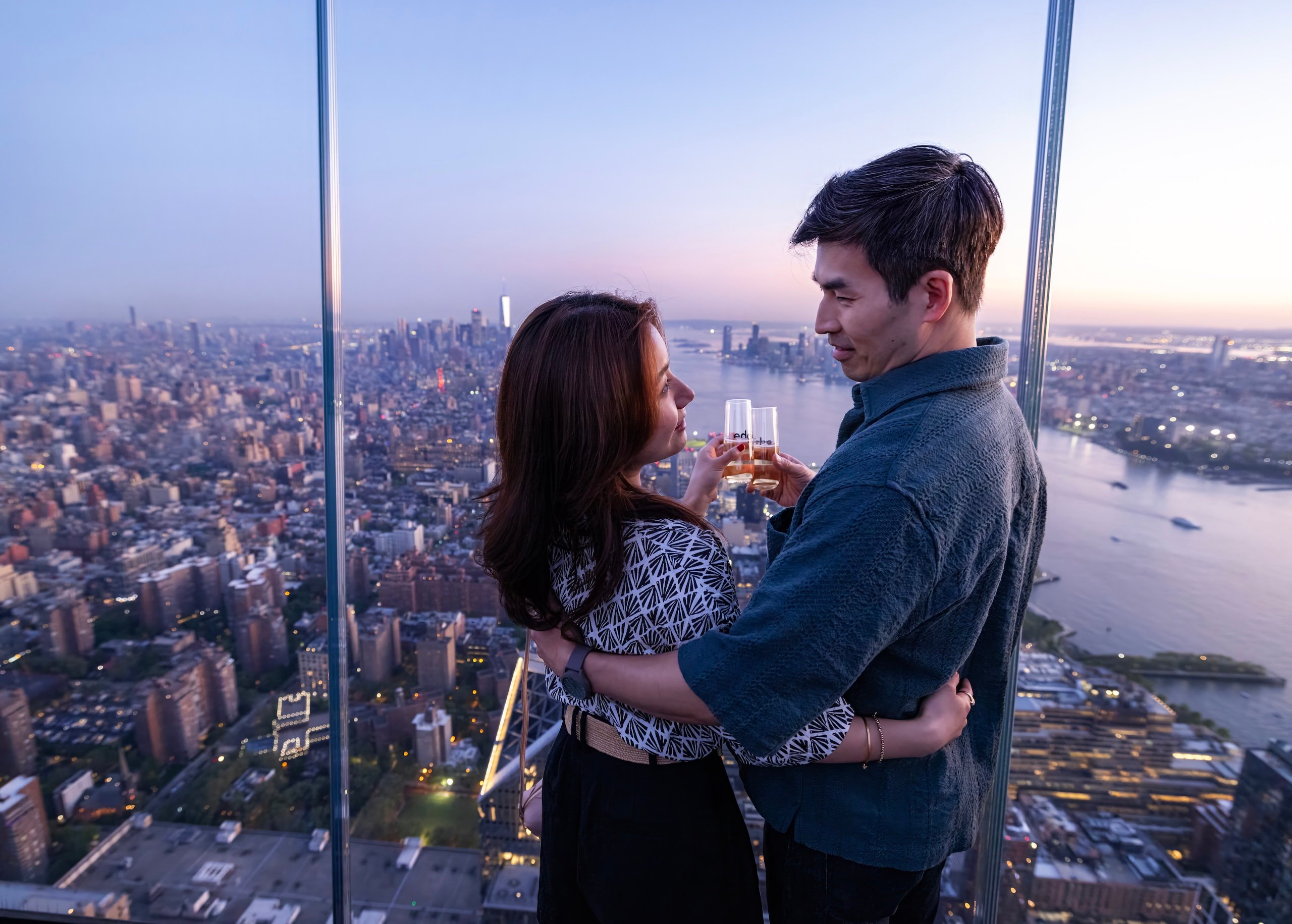 A couple clinks glasses of champagne overlooking a pink and purple sunset over New York city at The Edge