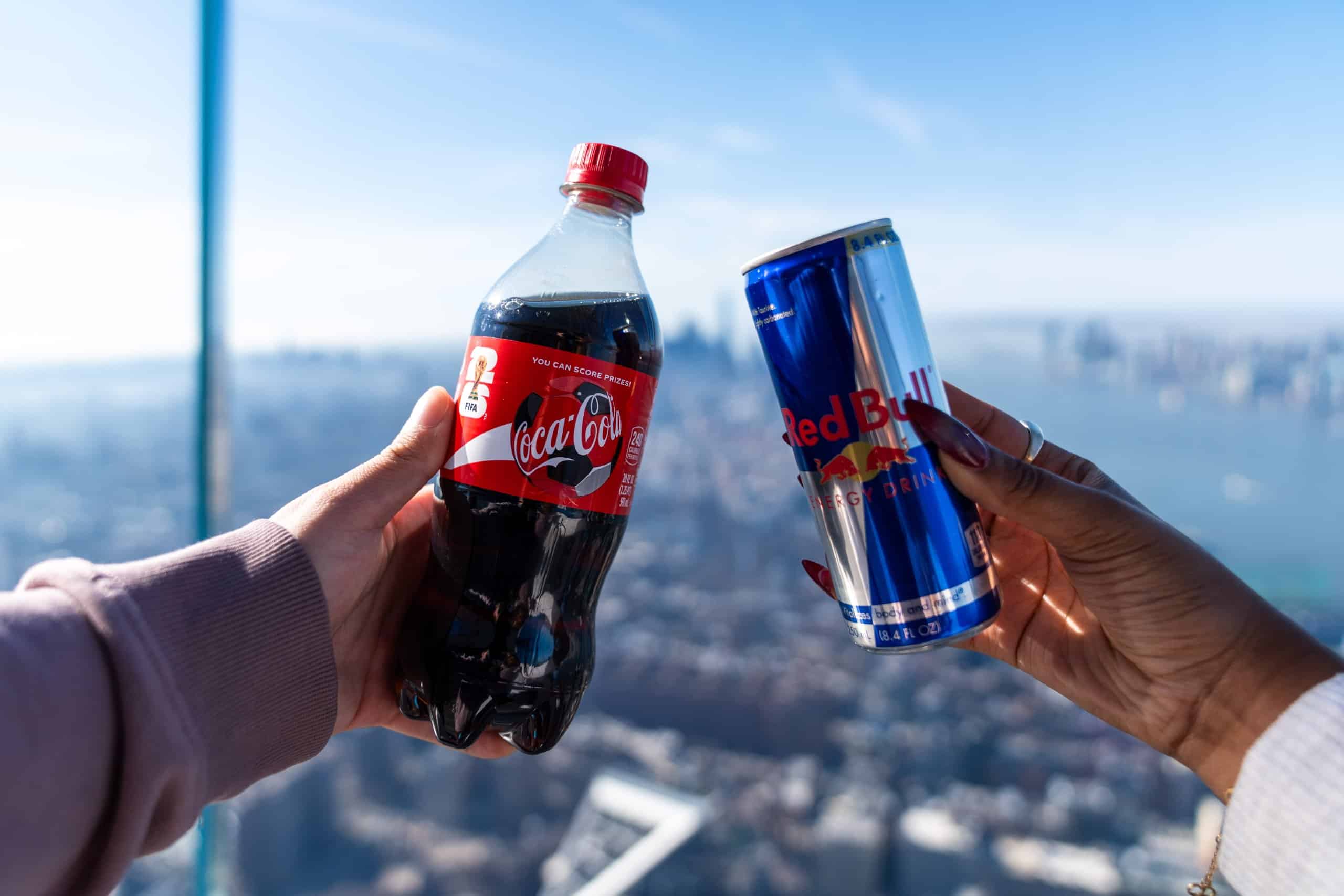 two hands holding a coca cola and redbull with the nyc skyline in the background