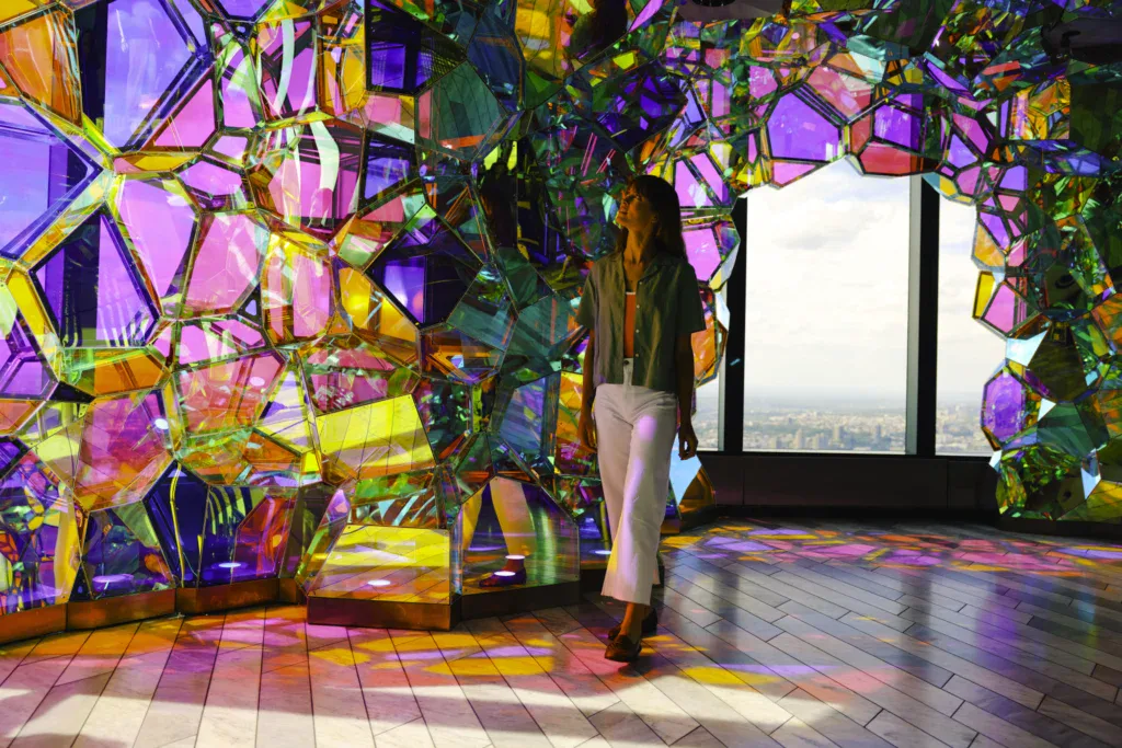 edge-crystal-cave-woman-skyline-view - Edge NYC - Edge NYC Woman in a green shirt and white pants stands among colorful mirrored panels, looking up with a city skyline visible through the window.