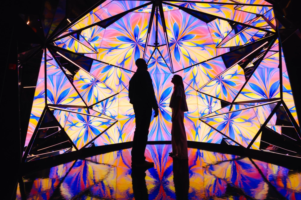 Edge NYC Light Art Installation - Edge NYC - Edge NYC Silhouetted man and woman stand before a vibrant geometric light installation with pink, yellow, and blue panels reflected on a glossy floor.