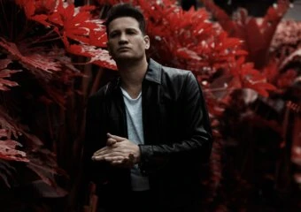 Portrait of a man in a black leather jacket standing among vibrant red foliage, looking off to the side.