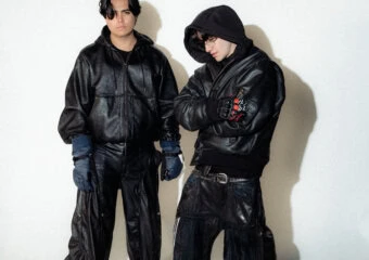 Two men in black leather outfits pose against a light backdrop, one with a hooded jacket and gloves on the left and the other with a hood and glasses on the right.